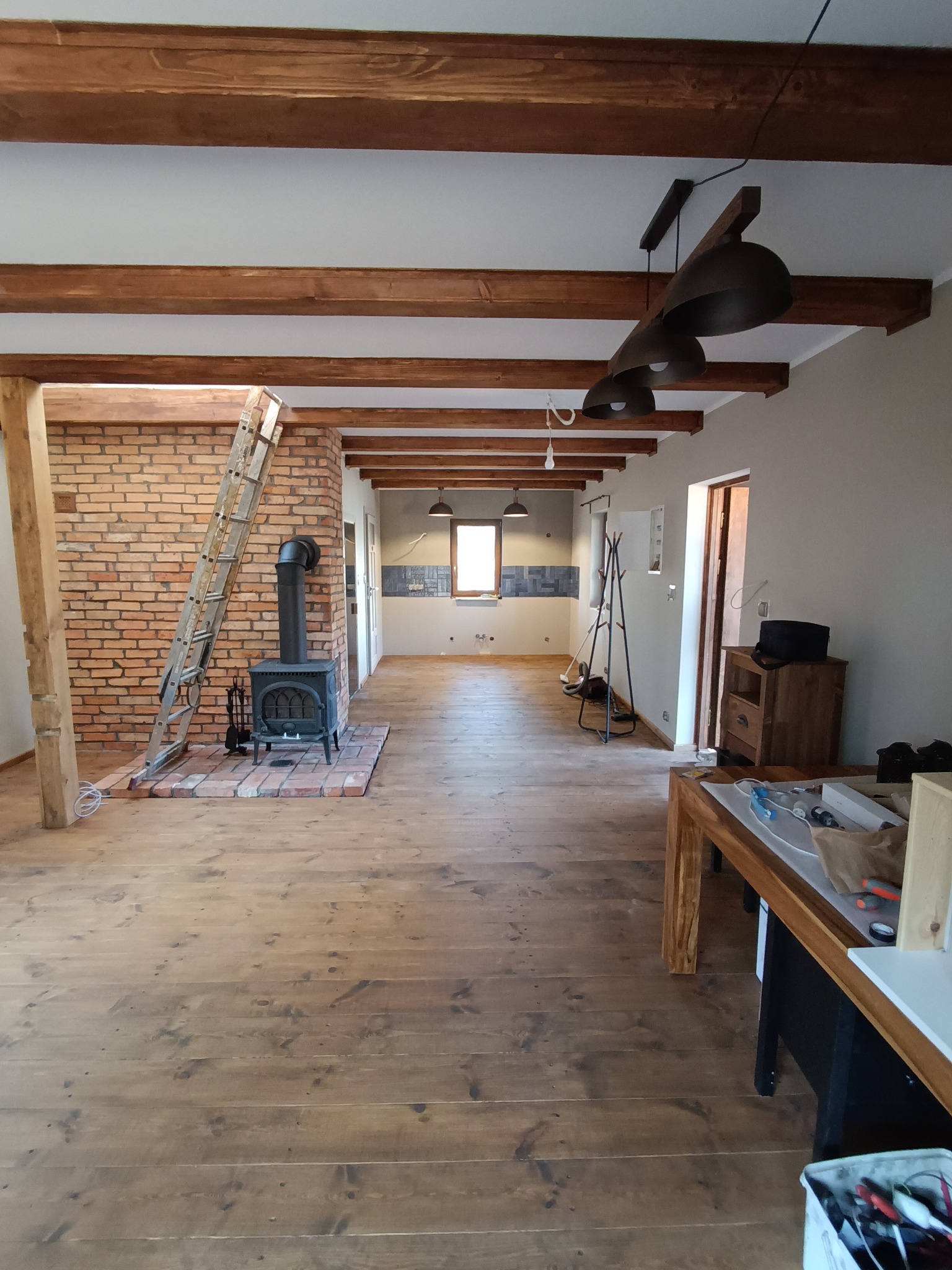 Rustykalne wnętrze z drewnianą podłogą, ceglaną ścianą i piecem. Widoczne drewniane belki stropowe i drabina oparta o ścianę. W głębi widoczna kuchnia.