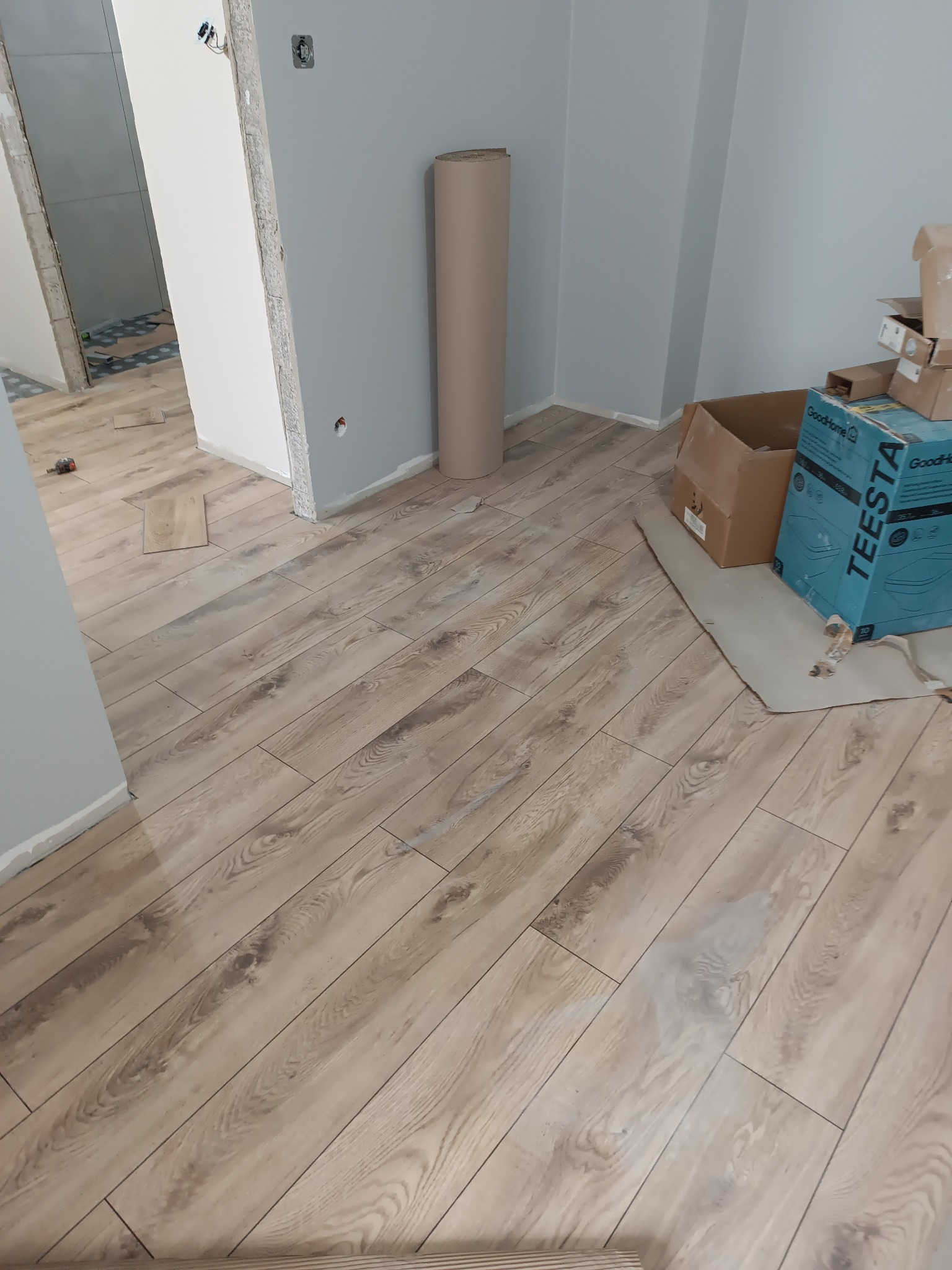 Jasne panele podłogowe ułożone w nowym pomieszczeniu. Widoczne kartony i rolka materiału pod ścianą. Fragment instalacji elektrycznej w ścianie.