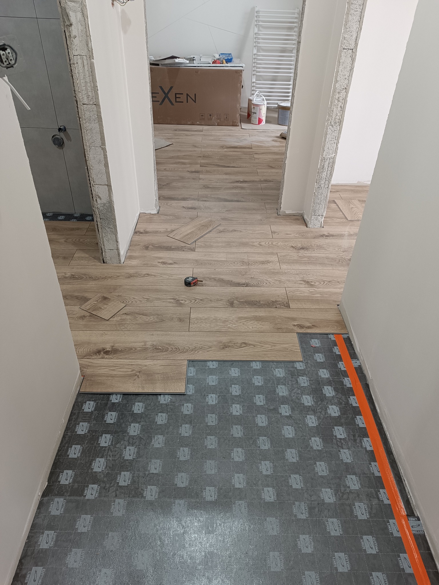 Układanie paneli podłogowych w korytarzu. Widoczna podkładka pod panele i częściowo ułożona podłoga z paneli imitujących drewno. Pomarańczowa taśma zabezpieczająca przy ścianie.