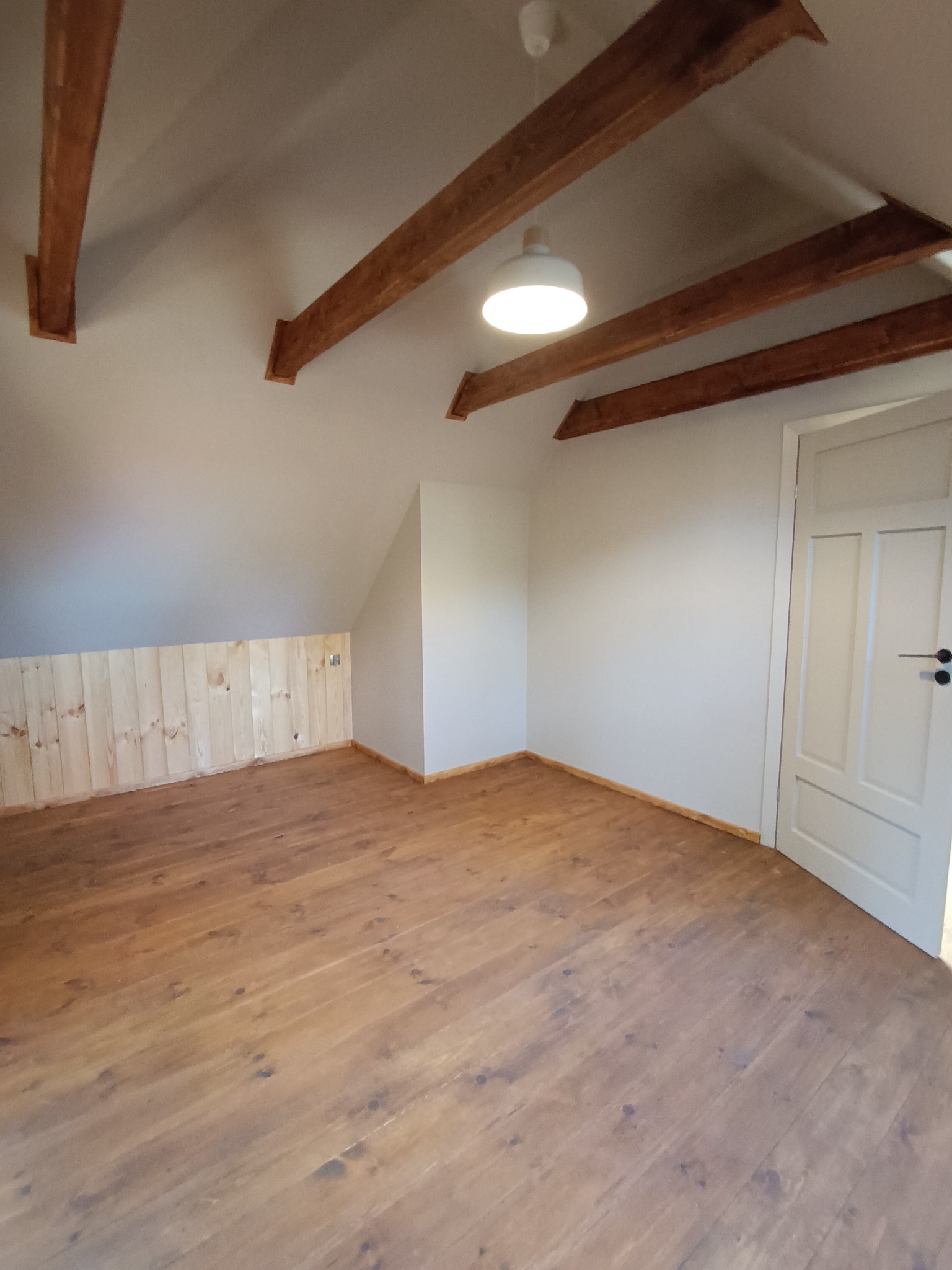 Jasne poddasze z drewnianą podłogą i boazerią. Widoczne drewniane belki stropowe i białe drzwi. Całość oświetlona centralnym plafonem.