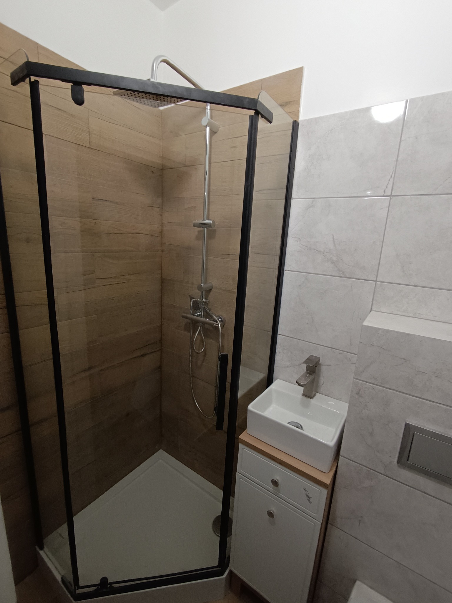 Nowoczesna kabina prysznicowa z czarną ramą i deszczownicą, obok umywalka nablatowa na szafce. Ściany wyłożone płytkami imitującymi drewno i marmur.