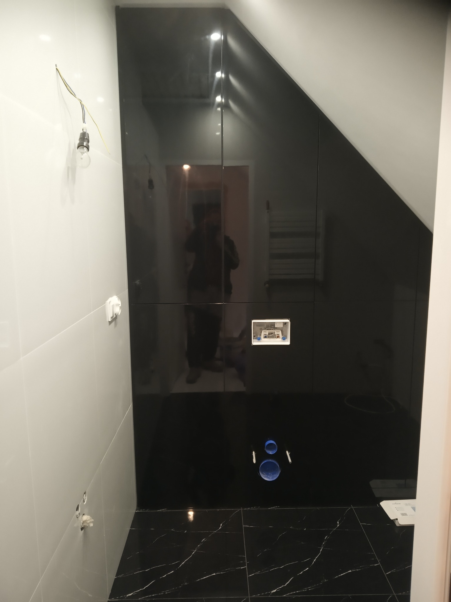 Wykończona łazienka w odcieniach bieli i czerni, z czarną ścianą z połyskiem i marmurową podłogą, widoczne przygotowane przyłącza wodne i elektryczne, surowa żarówka zwisająca z sufitu...