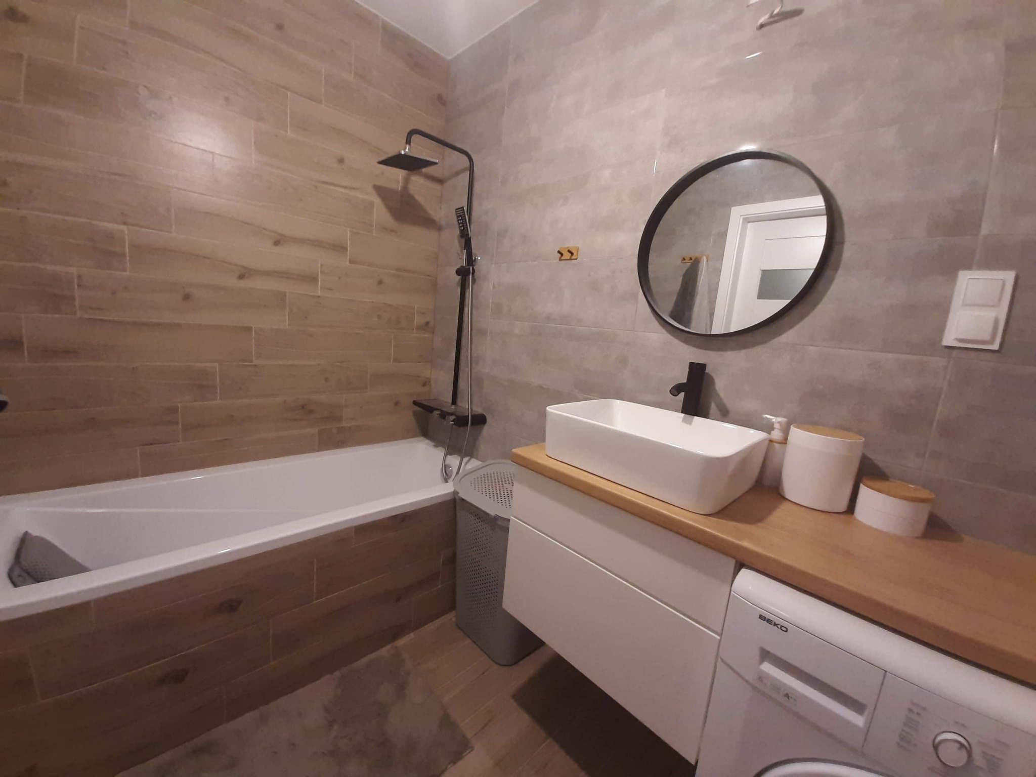Nowoczesna łazienka z wanną obłożoną drewnopodobnymi płytkami, czarną armaturą, okrągłym lustrem i białą umywalką nablatową na szafce z drewnianym blatem, pod którą znajduje się pralka Beko.