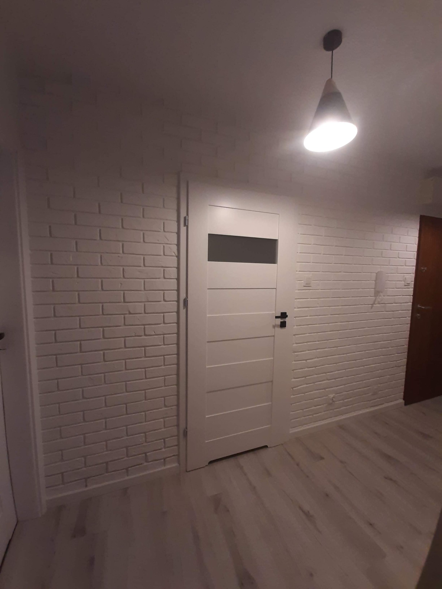 Wnętrze po odnowieniu: biała ściana z cegły, białe drzwi z poziomym przeszkleniem i panele podłogowe w odcieniach szarości, oświetlone nowoczesną lampą wiszącą.