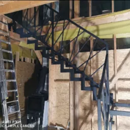 Konstrukcja schodów z czarnego metalu z geometrycznymi poręczami, widoczna drabina aluminiowa i piec w surowym wnętrzu domu w trakcie budowy.