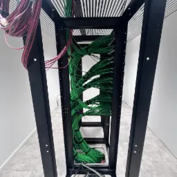 Wnętrze szafy rack z uporządkowanymi wiązkami kabli w kolorze zielonym i luźnymi kablami czarnymi na dole, widok z dołu, minimalistyczne białe ściany w tle.