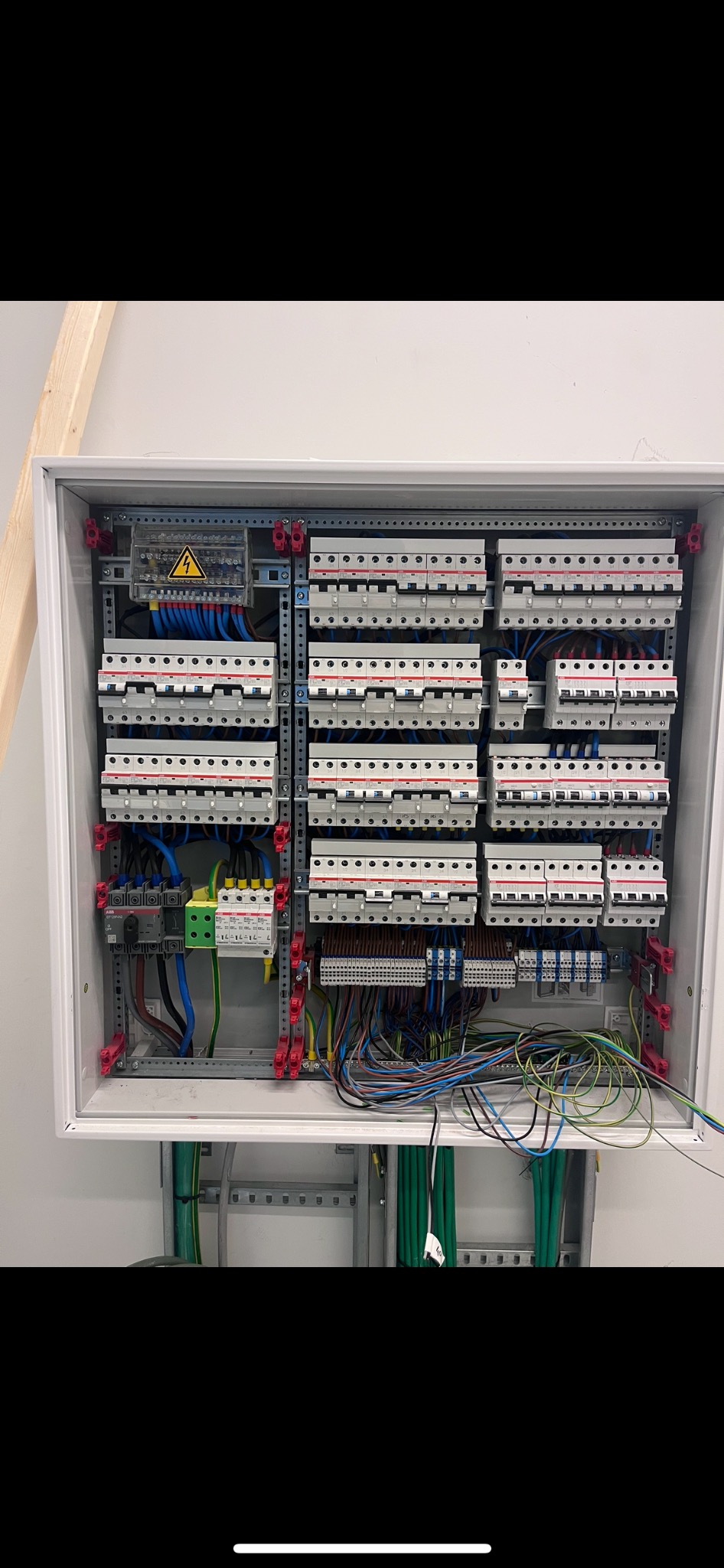 Rozdzielnica elektryczna z widocznymi bezpiecznikami, przewodami i elementami zabezpieczającymi, zamontowana w białej obudowie, z żółtym znakiem ostrzegawczym wysokiego napięcia w lewym górnym rogu.