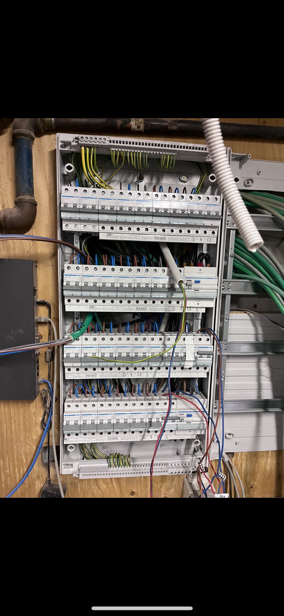 Otwarta skrzynka rozdzielcza z widocznymi bezpiecznikami, przewodami w różnych kolorach, zamontowana na drewnianej ścianie, obok rura instalacyjna i czarny moduł elektroniczny.