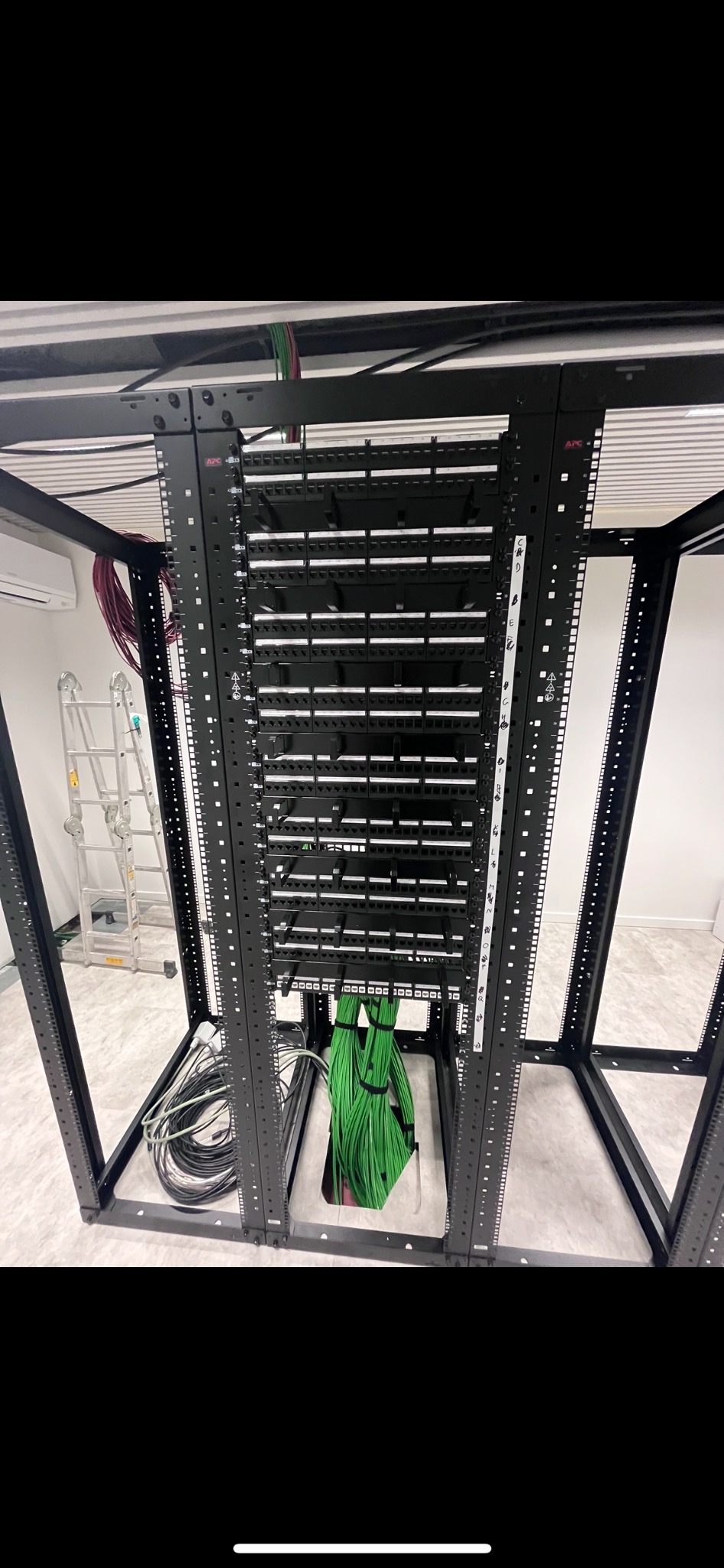 Widok z dołu na czarną szafę rack z panelami krosowymi i uporządkowanymi kablami w kolorze zielonym i czarnym, drabina aluminiowa oparta o ścianę w tle.