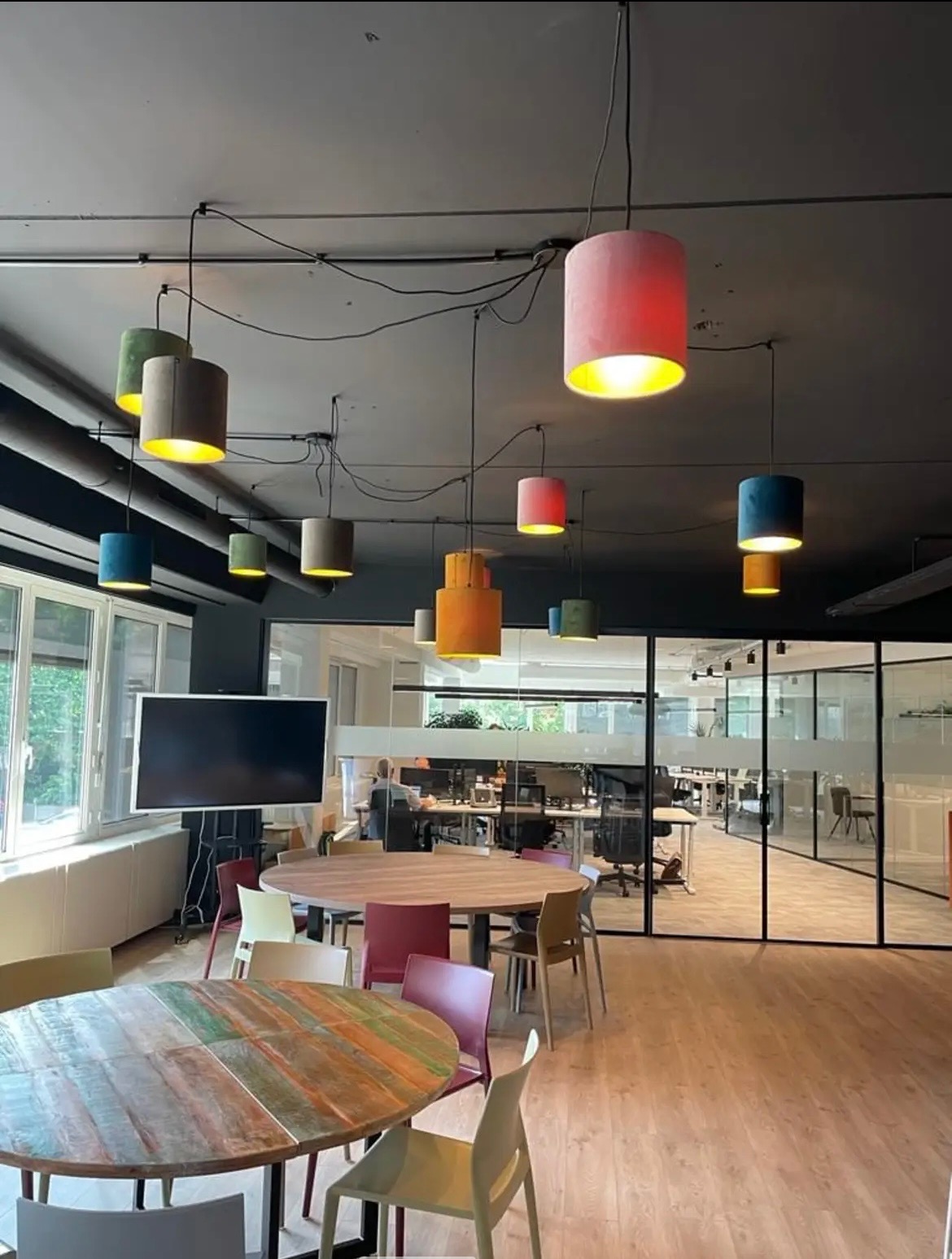 Wnętrze biura z różnokolorowymi, cylindrycznymi lampami wiszącymi nad okrągłymi stołami, widok z perspektywy podłogi, w tle przeszklone biura z pracownikami przy biurkach.