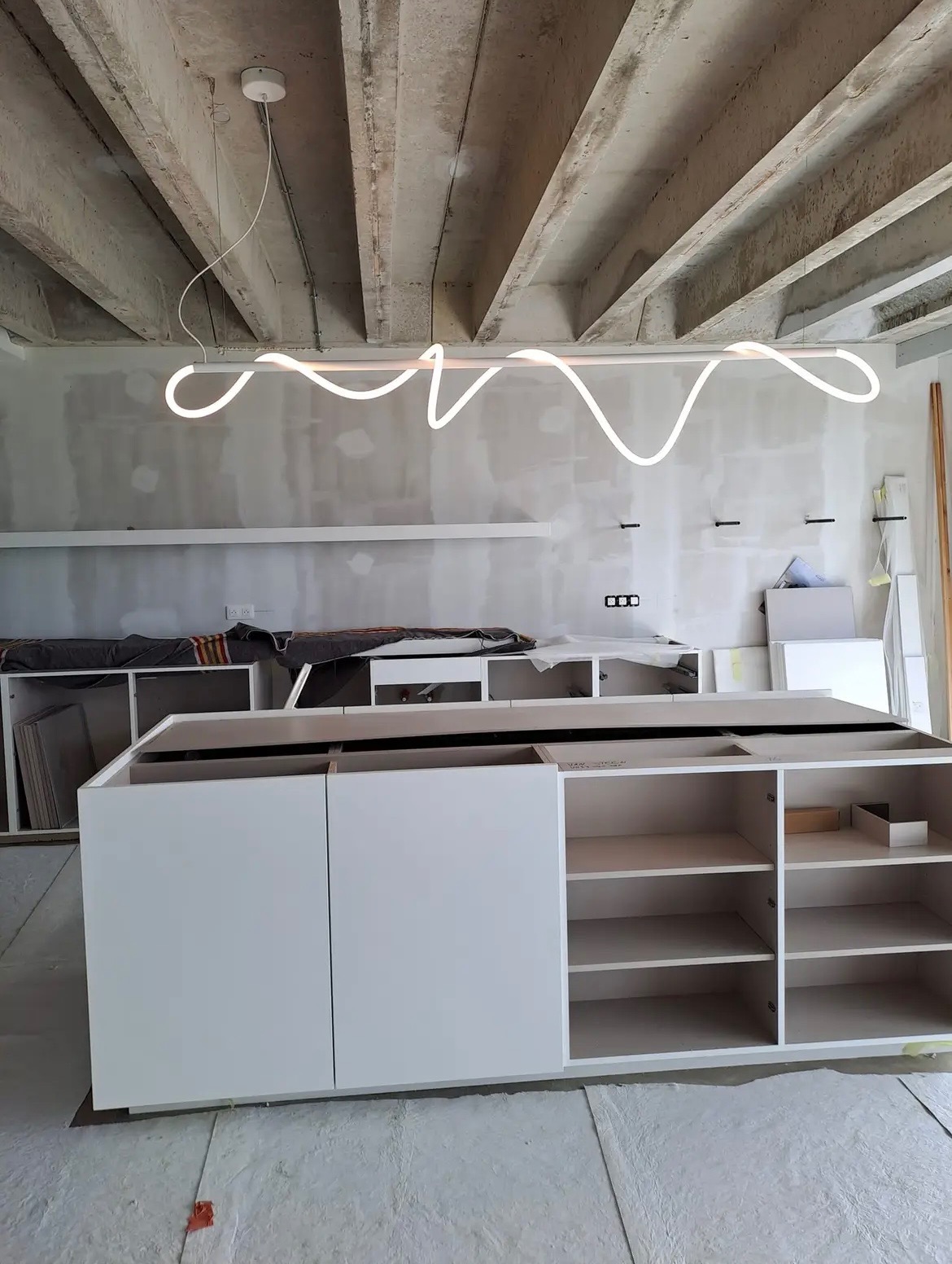 Wnętrze w trakcie remontu z surowym betonowym stropem, nowoczesną, falistą lampą LED i częściowo zmontowaną, białą zabudową meblową.