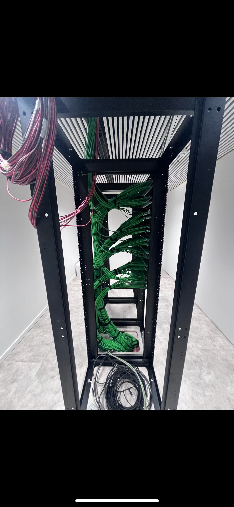 Wnętrze szafy rack z uporządkowanymi wiązkami kabli w kolorze zielonym i luźnymi kablami czarnymi na dole, widok z dołu, minimalistyczne białe ściany w tle.