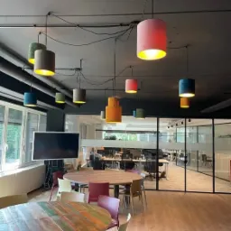 Wnętrze biura z drewnianymi stołami i krzesłami, oświetlone wiszącymi lampami w różnych kolorach i kształtach, z widocznymi przewodami instalacji elektrycznej na suficie.