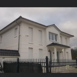 Dwupiętrowy dom z białymi ścianami i czarnym dachem, ogrodzony czarnym, kutym płotem. Widoczny czerwony czujnik alarmowy na elewacji.