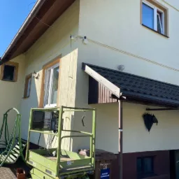 Dom jednorodzinny z zamontowanymi kamerami monitoringu, widoczne przewody i zielony podnośnik koszowy przy ścianie budynku.