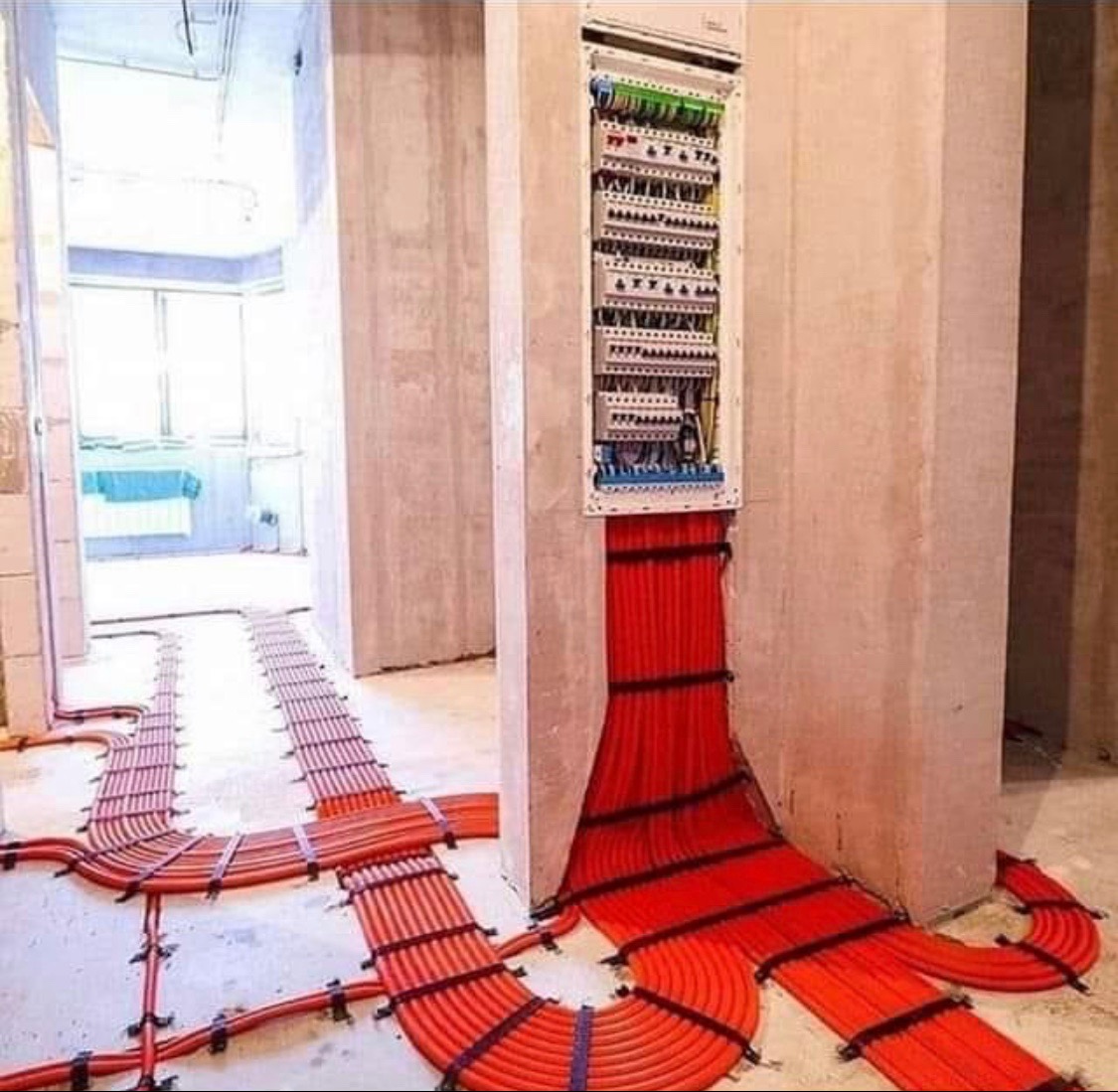 Rozdzielnica elektryczna z licznymi przewodami w pomarańczowych rurkach, ułożonymi równolegle i przymocowanymi czarnymi opaskami, prowadzącymi po podłodze w surowym wnętrzu budynku w trakcie prac...