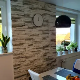 Ściana wykończona dekoracyjną okładziną imitującą kamień, z zegarem i roślinami doniczkowymi na parapecie, oświetlona wiszącą lampą w kształcie kuli, widok na stół z owocami.