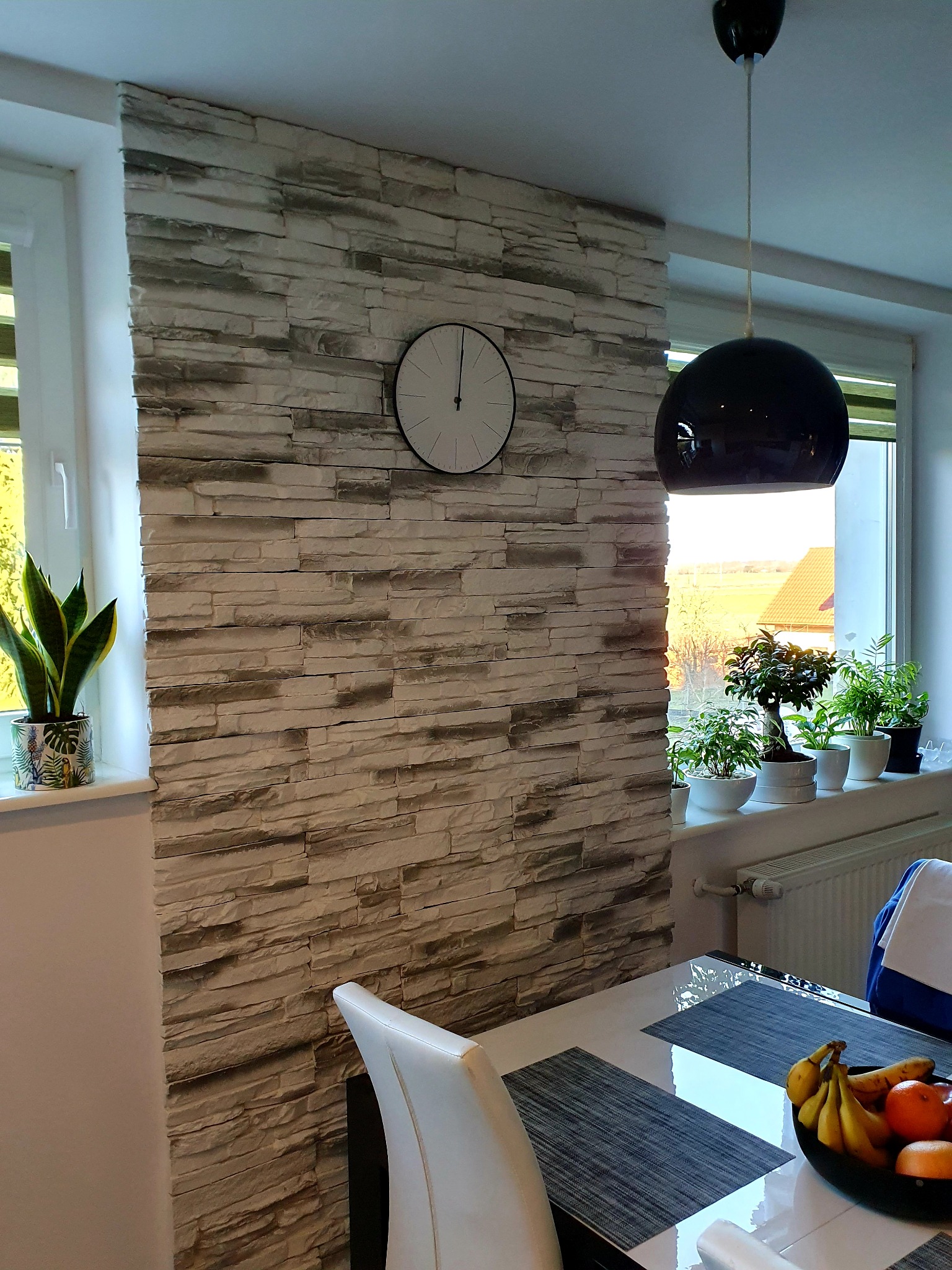 Ściana wykończona dekoracyjną okładziną imitującą kamień, z zegarem i roślinami doniczkowymi na parapecie, oświetlona wiszącą lampą w kształcie kuli, widok na stół z owocami.