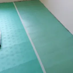 Zielona pianka pod panele, częściowo rozłożona na podłodze w pokoju, z białą taśmą wyznaczającą granicę ułożonej części.