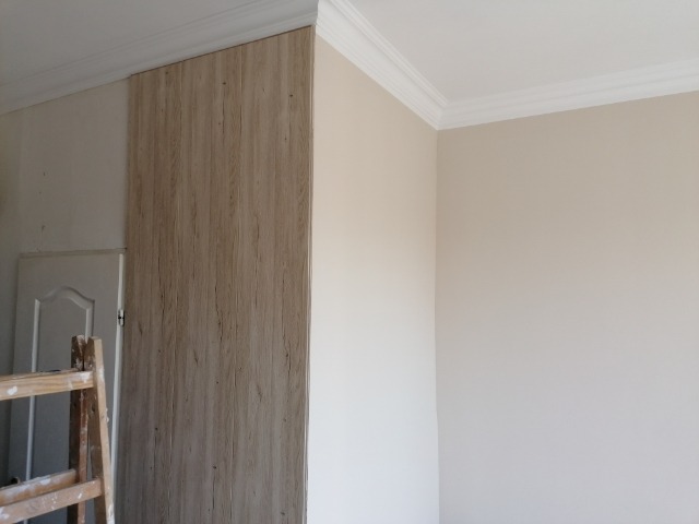 Ściana wykończona panelami imitującymi drewno, częściowo pomalowana na beżowo, widoczna drabina malarska i białe listwy przysufitowe.