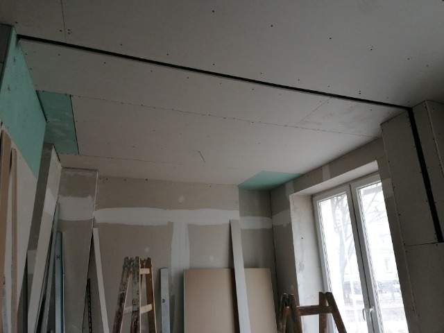 Montaż sufitu podwieszanego z płyt kartonowo-gipsowych, widoczne profile i płyty w trakcie instalacji, drabina oparta o ścianę, surowy stan wykończenia.