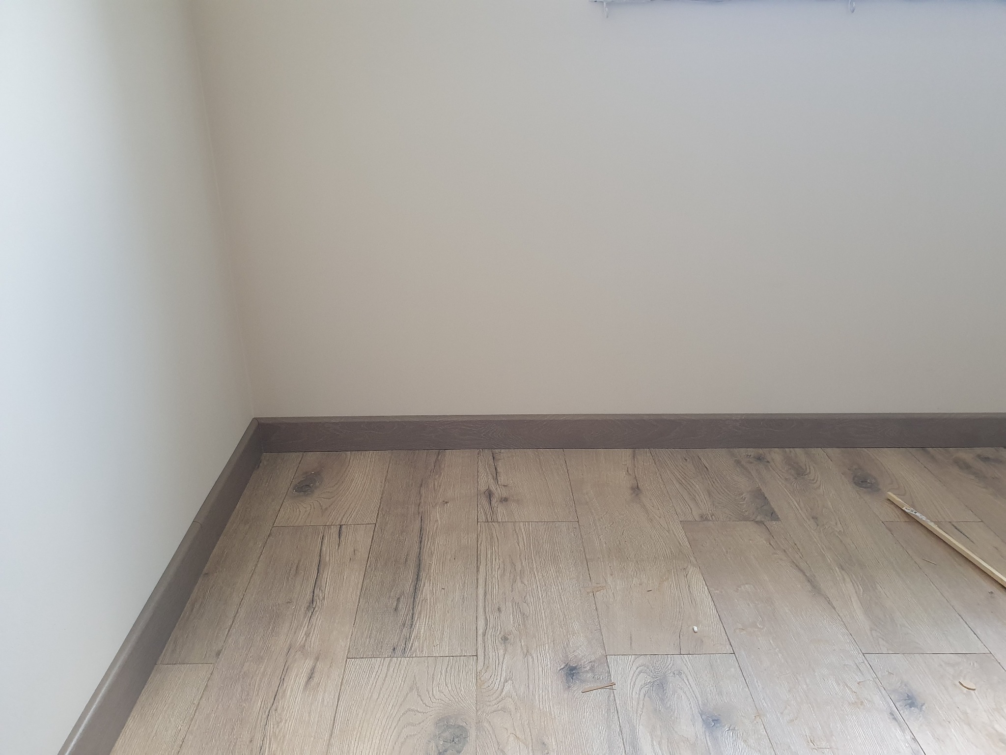 Fragment podłogi z jasnych paneli z widocznymi ciemnymi słojami, przy ścianie zamontowana szara listwa przypodłogowa, u góry kadru widoczny fragment białej ściany.