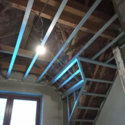 Metalowy stelaż pod sufitem na poddaszu, widoczna izolacja, okno w tle.