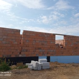 Kompleksowa obsługa inwestycji PP - Wznoszone ściany domu z czerwonej cegły ceramicznej, widoczne bloczki fundamentowe i ocieplenie fundamentów niebieskim styropianem, na drugim planie widoczny pracownik budowlany.