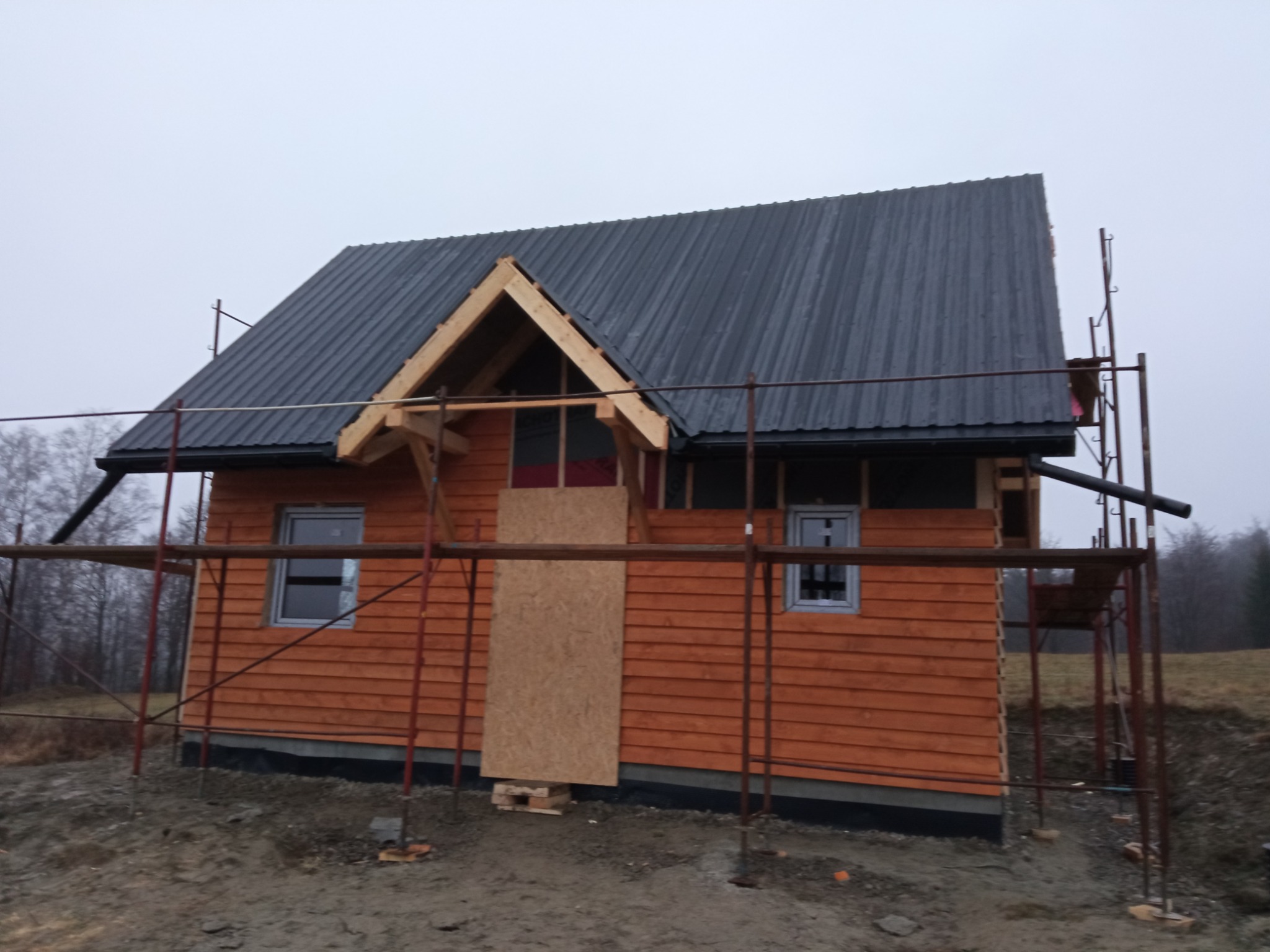 Surowy, drewniany dom w trakcie budowy, z widocznymi rusztowaniami, deskami elewacyjnymi w odcieniach brązu i szarą, metalową blachodachówką na dachu. Jedno okno zamaskowane płytą OSB.