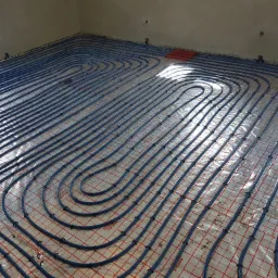 Nisko-temperaturowe ogrzewanie podłogowe z niebieskich rur fi 16mm ułożonych na folii z czerwoną siatką, widoczne uchwyty mocujące i fragmenty czerwonych rur przy ścianie, surowy stan wykończenia...