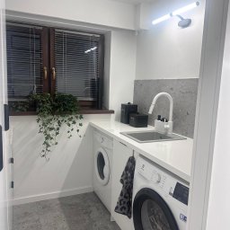 Meble na wymiar AG - Biała zabudowa pralni z pralkami, zlewem i blatem pod oknem z roletami. Na blacie doniczka z bluszczem i czarne pojemniki. Szara płytka nad zlewem, szara podłoga.