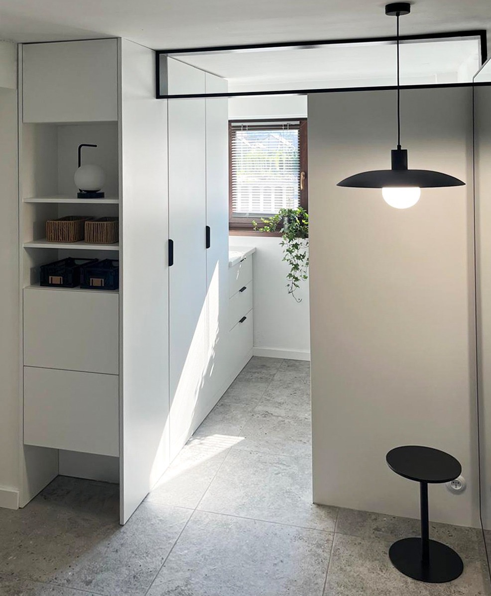 Biała szafa wnękowa z półkami i szafkami, czarne uchwyty, oświetlenie punktowe, szara podłoga, minimalistyczny design, roślina w oknie, nowoczesne wnętrze.