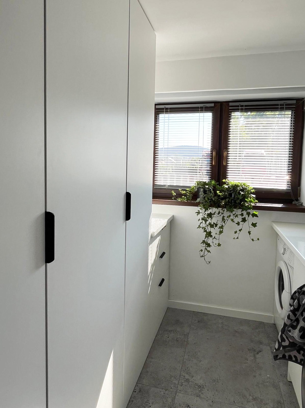 Biała szafa z czarnymi uchwytami w jasnym pomieszczeniu z oknem, na parapecie roślina. Widoczna pralka i szara podłoga. Minimalistyczny design.