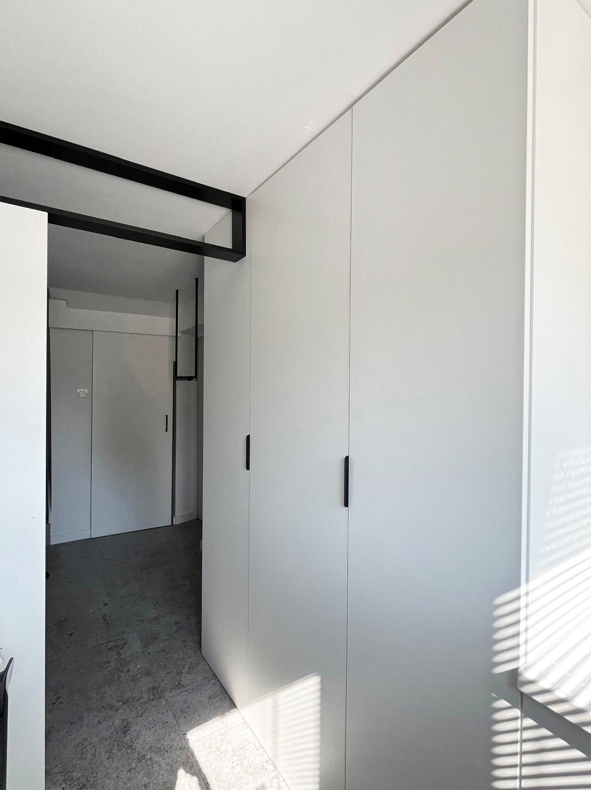 Minimalistyczna, biała szafa wnękowa z czarnymi, podłużnymi uchwytami, w jasnym pomieszczeniu z szarym kamieniem na podłodze i czarną ramą pod sufitem.