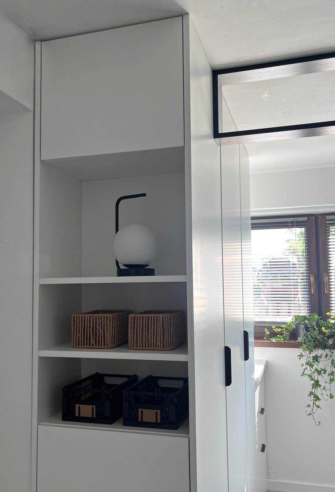 Biała szafa wnękowa z półkami, na których stoją dekoracyjne kosze i lampka. Widok na okno z roletami i rośliną doniczkową. Minimalistyczny design.