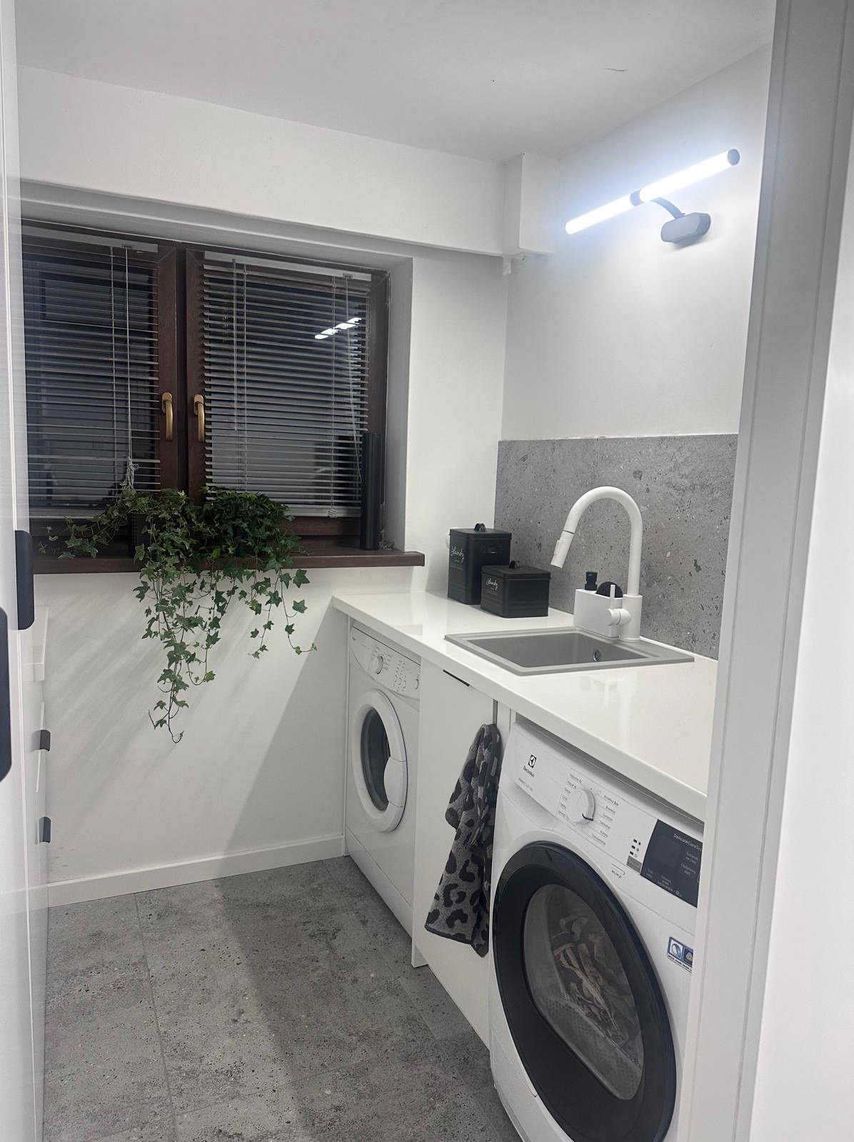Biała zabudowa pralni z pralkami, zlewem i blatem pod oknem z roletami. Na blacie doniczka z bluszczem i czarne pojemniki. Szara płytka nad zlewem, szara podłoga.
