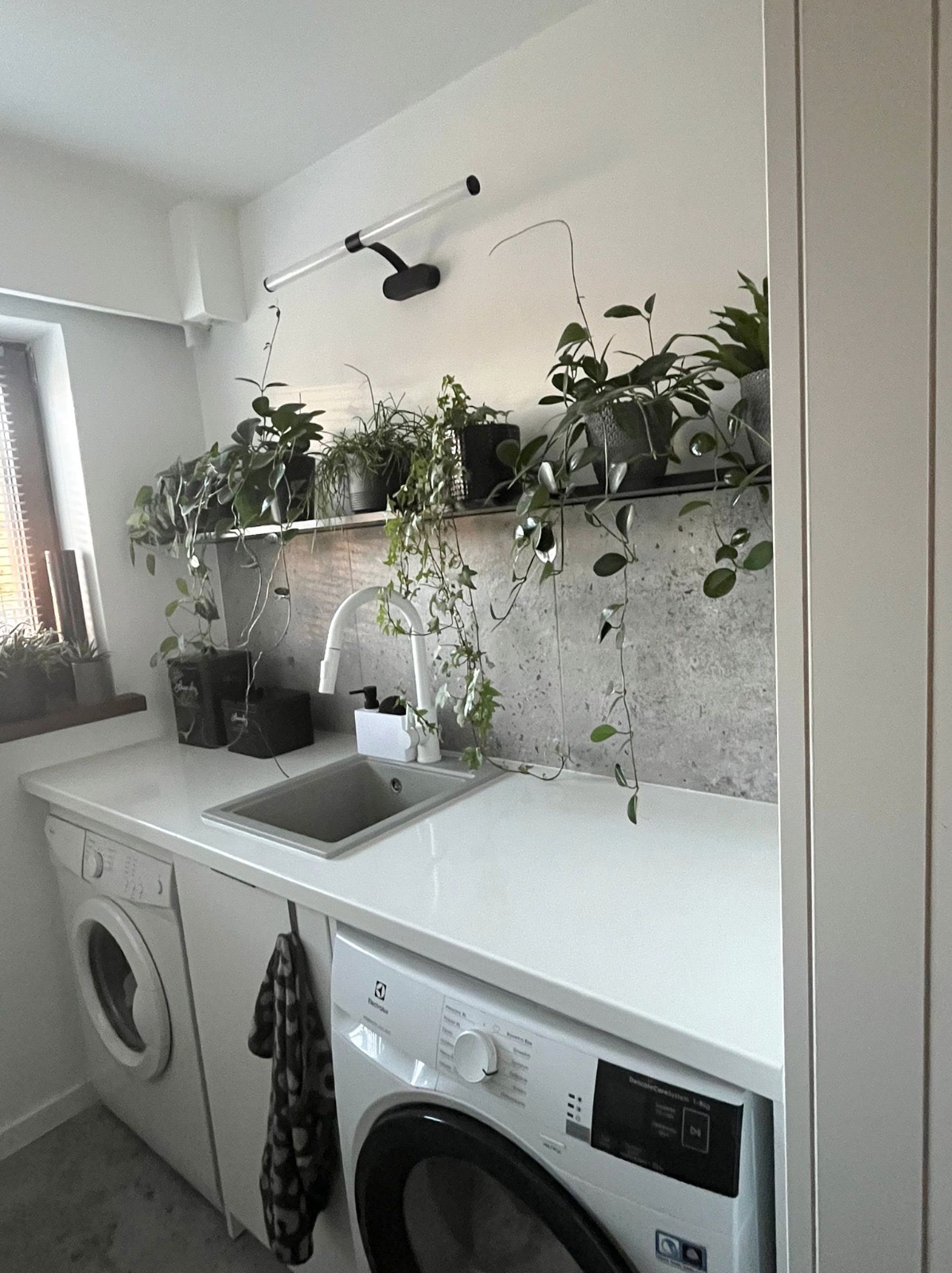 Biały blat z szarą umywalką i baterią, pod nim pralka Electrolux. Nad blatem półka z roślinami doniczkowymi, oświetlona lampą. Minimalistyczny wystrój wnętrza.