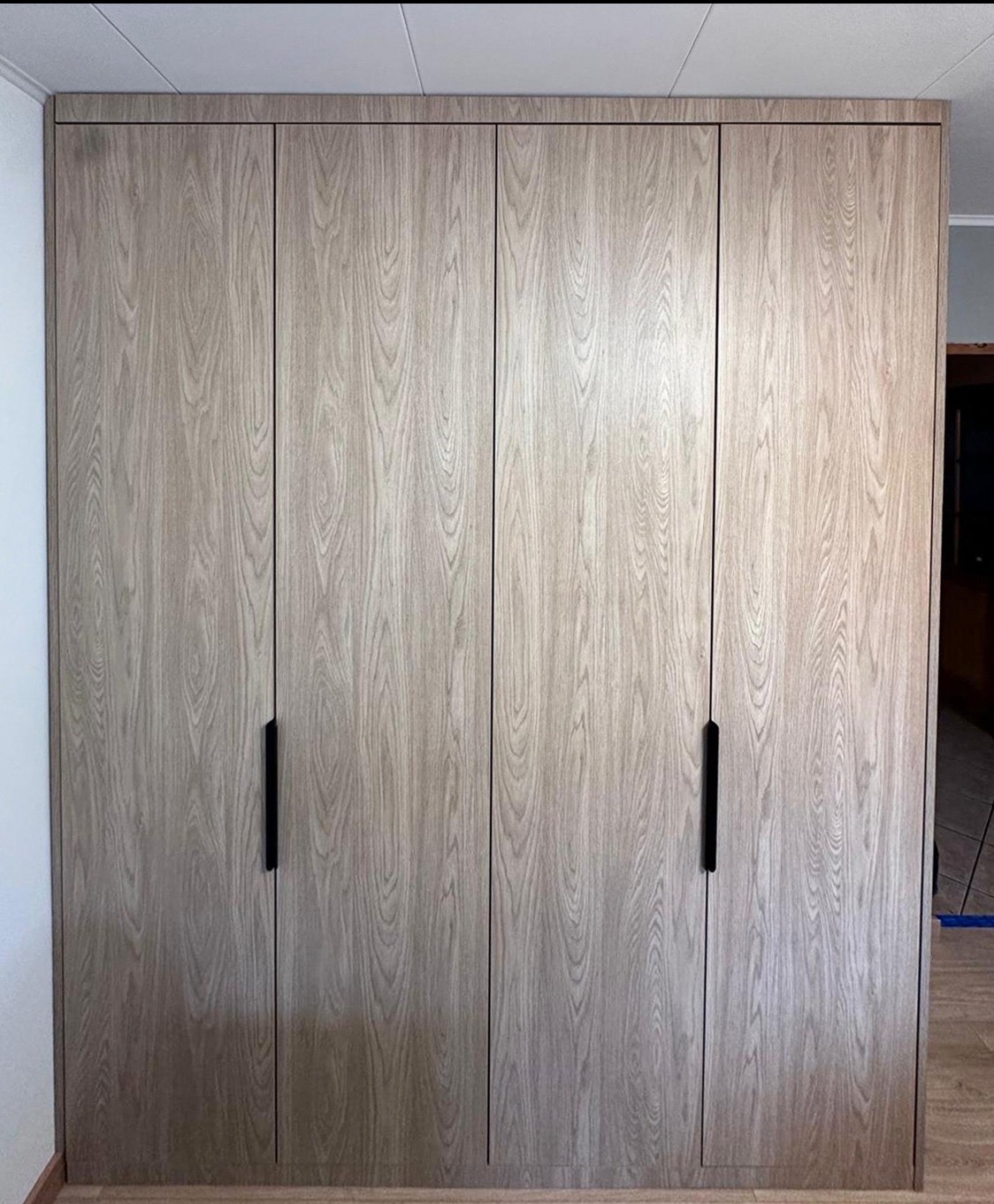 Czterodrzwiowa szafa wnękowa z jasnego drewna z ciemnymi, minimalistycznymi uchwytami, dopasowana do wnętrza, widoczna na tle ściany i podłogi.