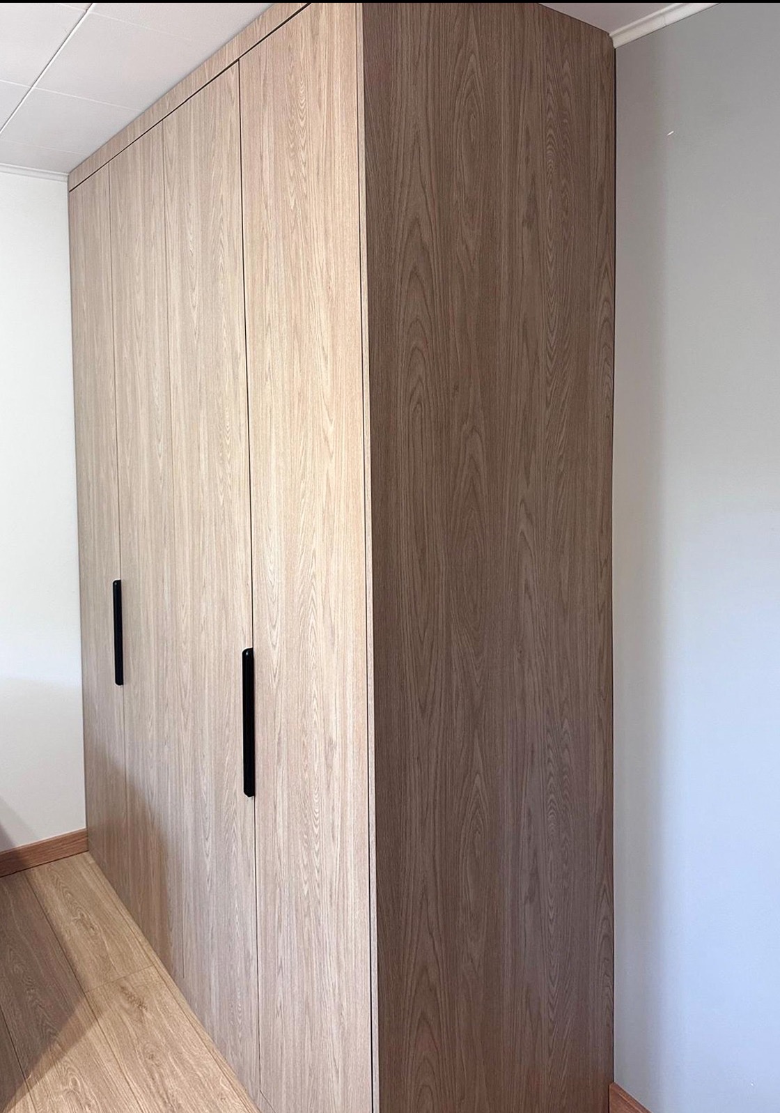 Jasna, drewnopodobna szafa wnękowa z czarnymi uchwytami, dopasowana do podłogi. Widok z boku, ukazujący pełną wysokość i zabudowę od podłogi do sufitu.
