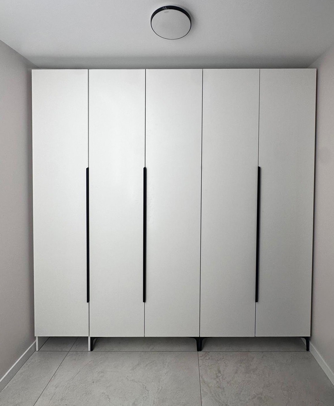 Biała, minimalistyczna szafa z czarnymi, długimi uchwytami, ustawiona na niskich nóżkach. Widoczna lampa sufitowa i płytki podłogowe w odcieniach szarości.