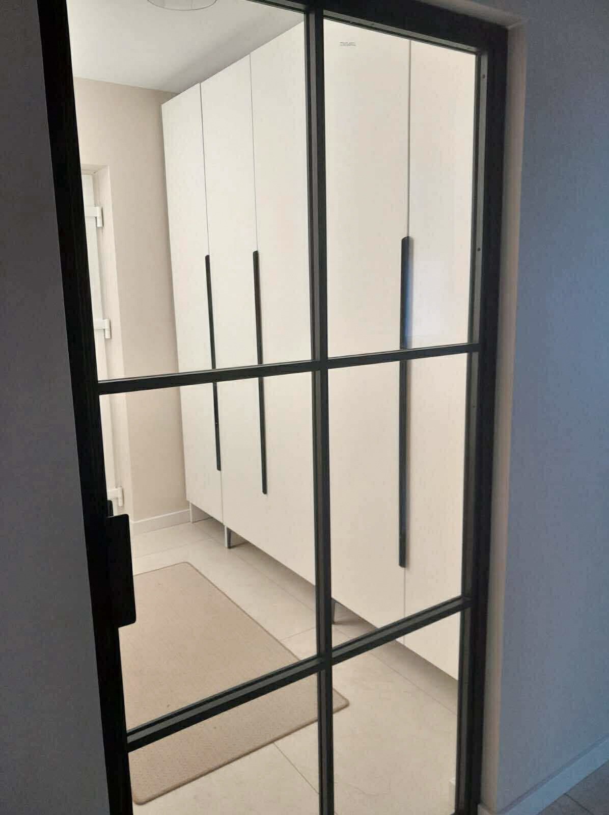 Biała szafa na wymiar z czarnymi, długimi uchwytami, widoczna przez przeszklone drzwi z czarną ramą. W tle beżowy dywanik i jasna podłoga.