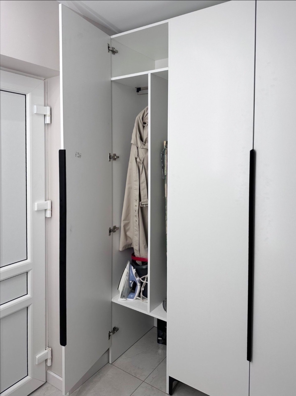 Biała szafa wnękowa z czarnymi uchwytami, otwarte drzwi ukazują półki z ubraniami i żelazkiem. Widoczny fragment drzwi wejściowych.
