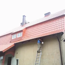 Pracownik na drabinie maluje elewację budynku z czerwoną dachówką, widoczne okna i komin