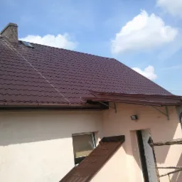 Brązowy dach z blachodachówki, widoczny komin, rynny i fragment elewacji budynku w jasnym odcieniu, na tle błękitnego nieba z chmurami.