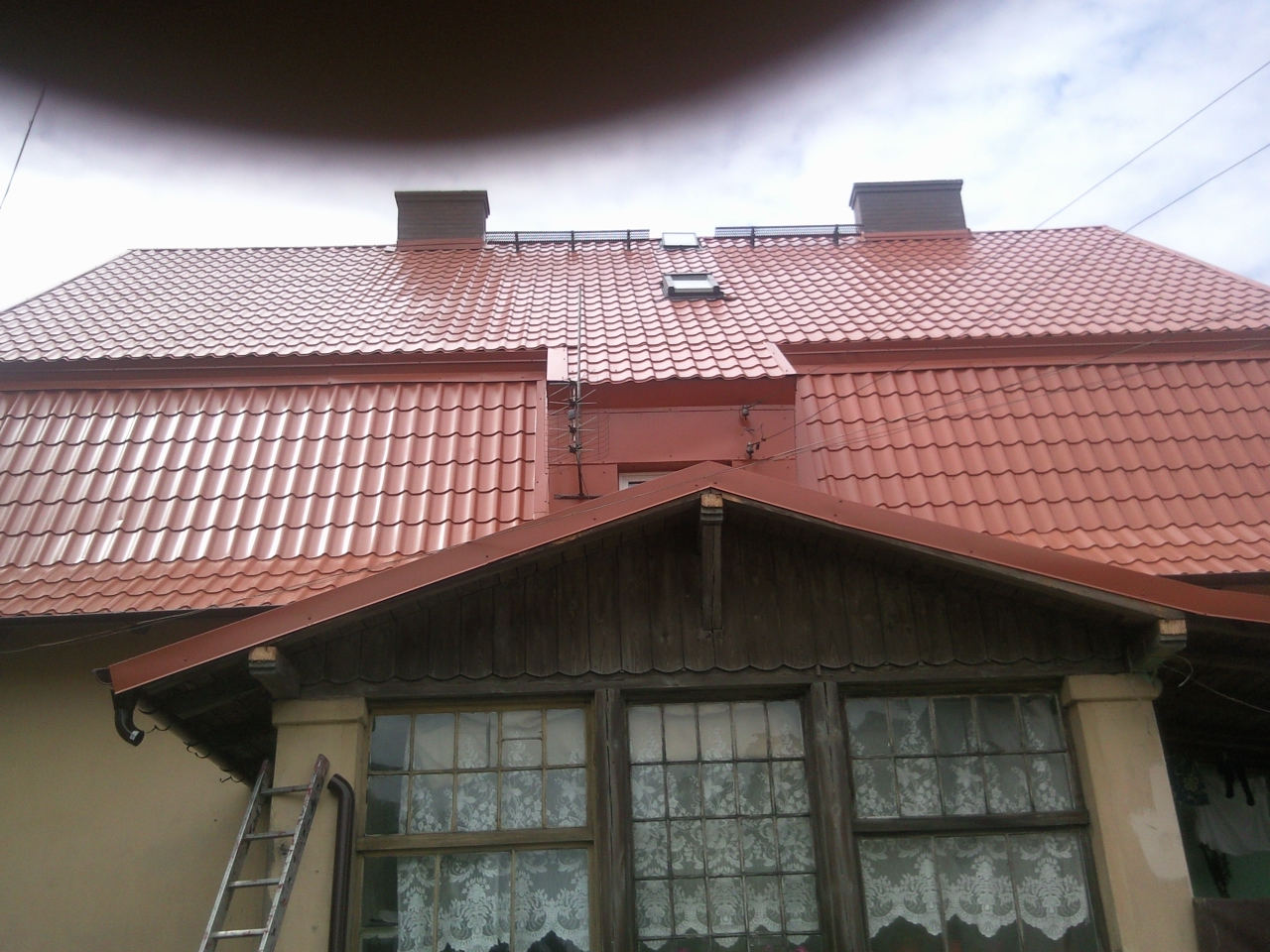 Dom z nowym, błyszczącym dachem z dachówki ceramicznej w odcieniach czerwieni, widoczne kominy i okno dachowe, drabina oparta o ścianę budynku.