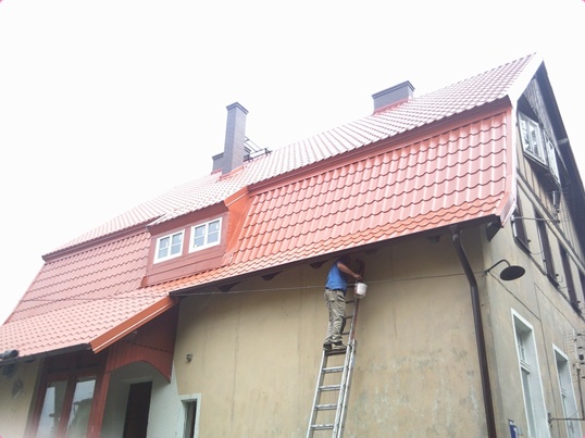 Pracownik na drabinie maluje elewację budynku z czerwoną dachówką, widoczne okna i komin