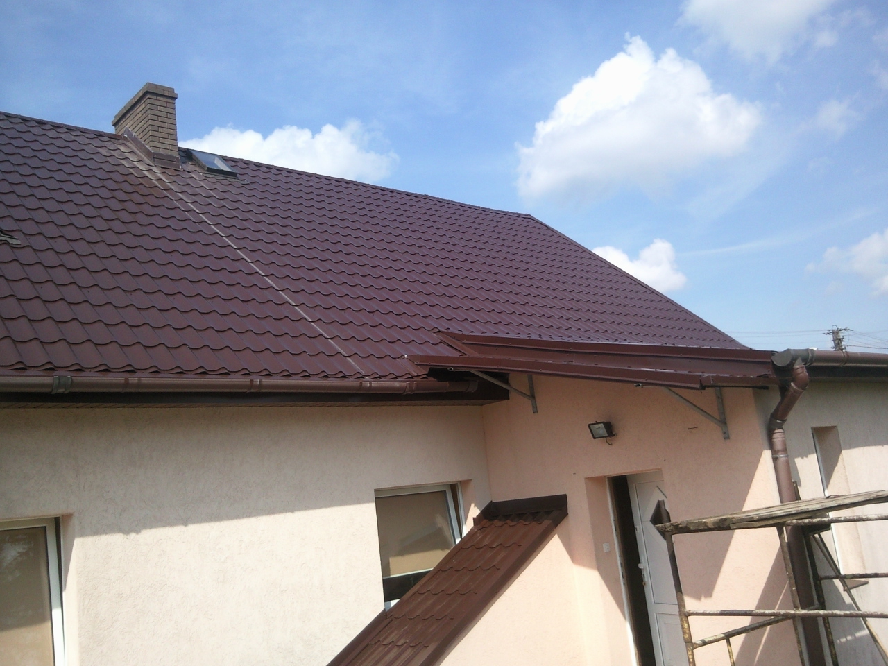 Brązowy dach z blachodachówki, widoczny komin, rynny i fragment elewacji budynku w jasnym odcieniu, na tle błękitnego nieba z chmurami.