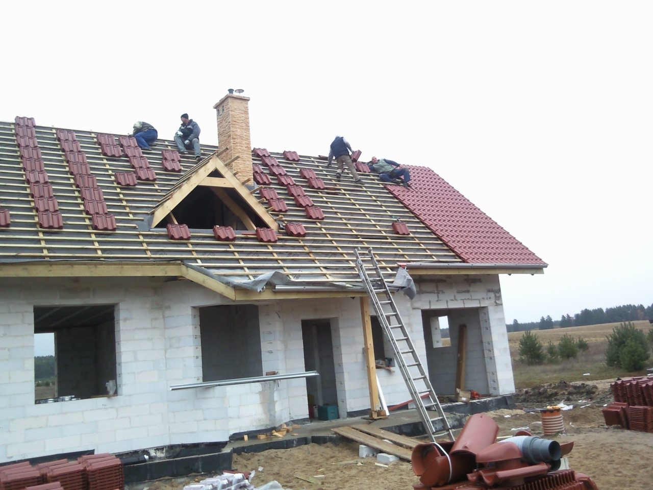 Ekipa dekarzy układa czerwoną dachówkę na drewnianej konstrukcji dachu domu w trakcie budowy, widoczny komin z cegły, stosy dachówek na ziemi i aluminiowa drabina oparta o budynek z pustaków.