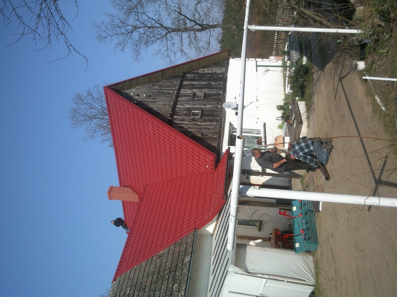 Czerwony dach z blachodachówki na starym domu, dwóch mężczyzn przy gruncie przycina metalową rurę, widoczne narzędzia i skrzynki narzędziowe.