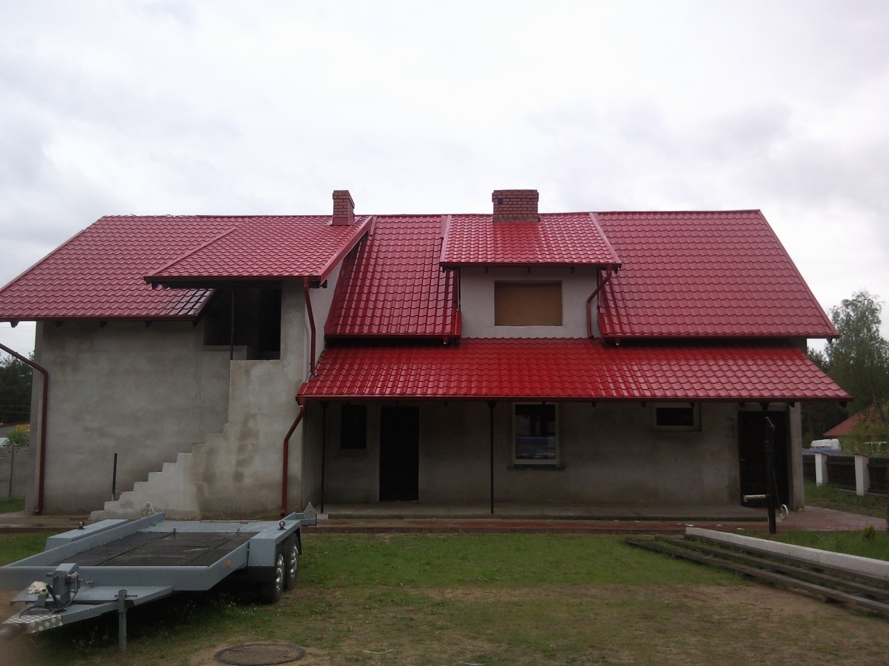 Dom w stanie surowym otwartym w Człuchowie z nowym, czerwonym dachem z blachodachówki, widoczna przyczepka samochodowa.